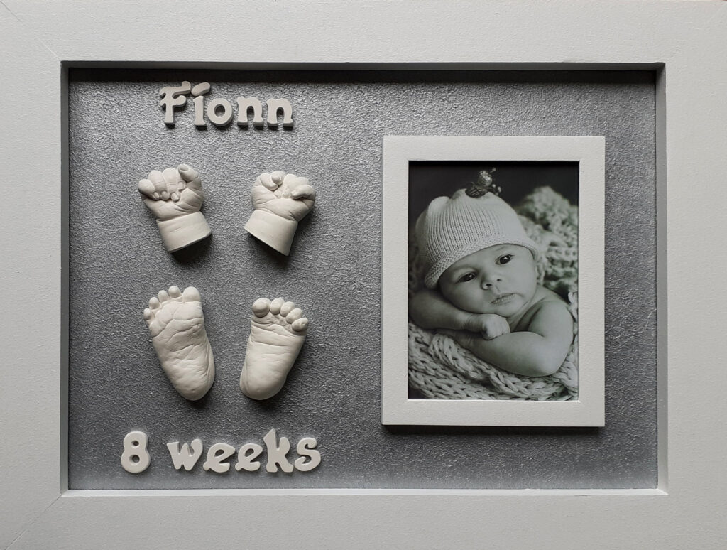silver Framed artwork showing white casts of a baby’s hands and feet on a grey background, alongside a black-and-white photo of a baby.